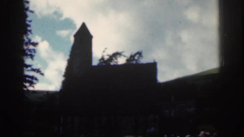 1961 Dublin-area home-movie frame with a church tower and roofline silhouetted against bright clouds, with trees and hillside nearby.
