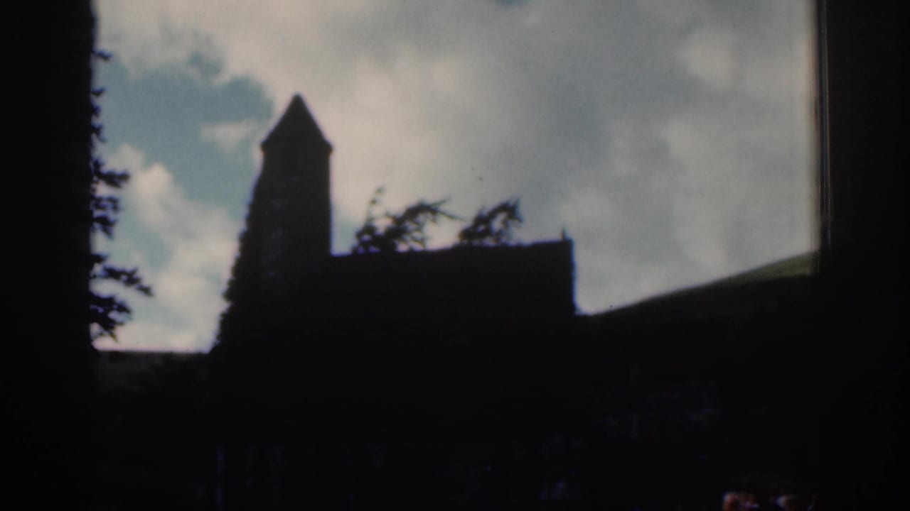 1961 Dublin-area 8mm view of a church tower and dark roofline silhouetted against clouds, with trees and a low hillside in the background.