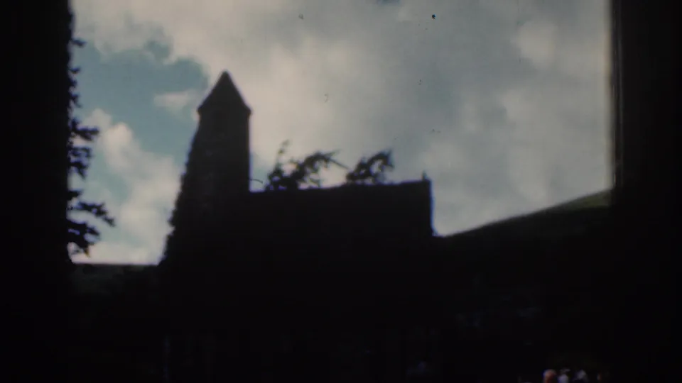 1961 Dublin-area 8mm frame with a silhouetted stone church tower, trees, hillside, and cloudy sky.