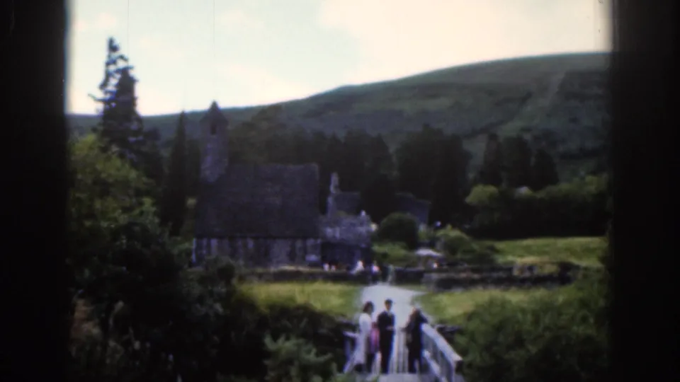 1961 Dublin-area home-movie frame showing visitors on a bridge with a stone church, trees, and hills beyond.