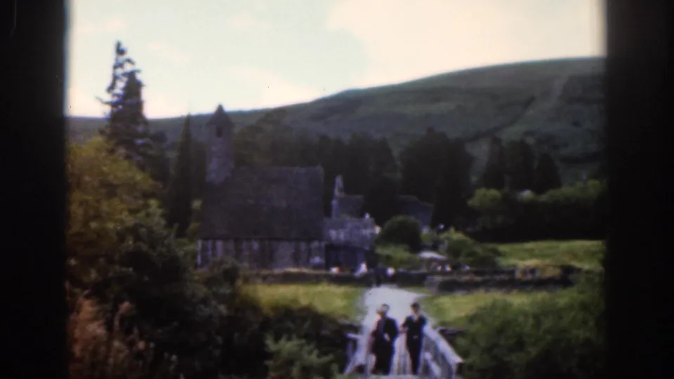 1961 Dublin-area 8mm frame with three visitors on a footbridge leading toward a stone church and hillside.