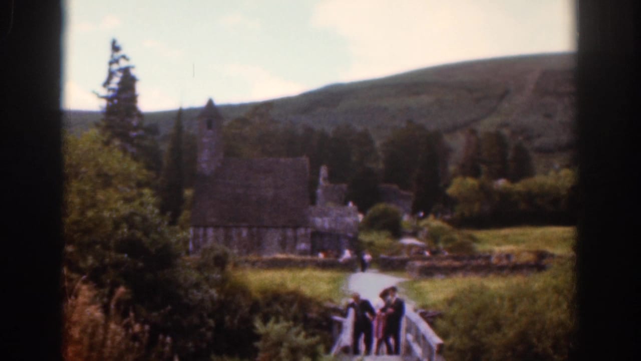 1961 Dublin-area home-movie view of tourists crossing a wooden bridge toward stone estate buildings and a small chapel-like tower.