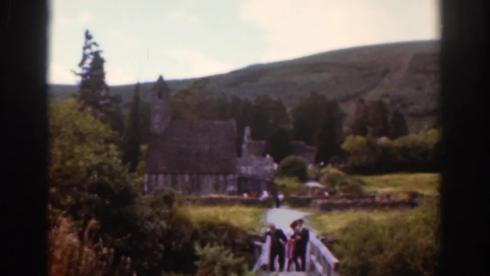 1961 Dublin-area home-movie view of tourists crossing a wooden bridge toward stone estate buildings and a small chapel-like tower.