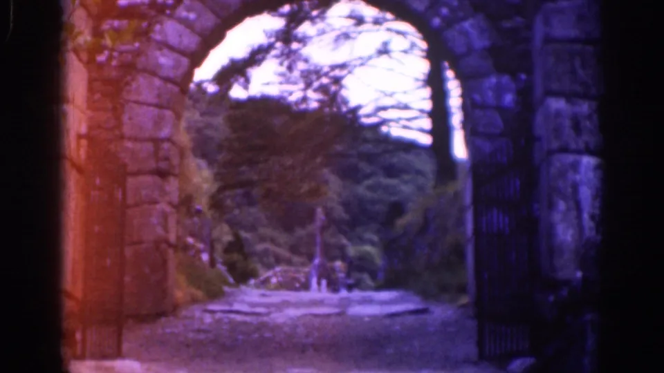 Visitor-shot 8mm view of a stone archway and iron gate looking toward a wooded path in Ireland, 1961.