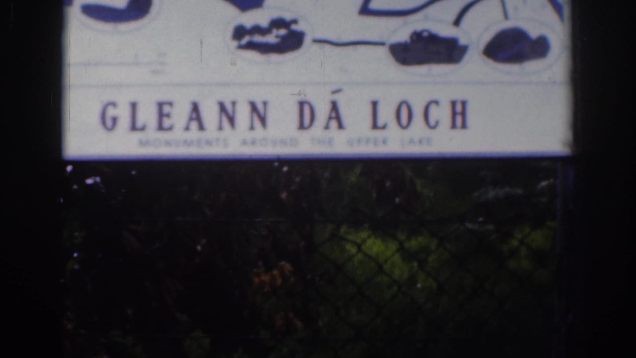 1961 8mm view of the Gléann Dá Loch sign, softly focused with aged color and dark foliage underneath.
