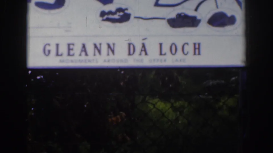 1961 8mm view of the Gléann Dá Loch sign, softly focused with aged color and dark foliage underneath.