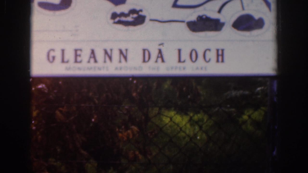 1961 8mm frame of a Gleann Da Loch sign viewed through chain-link fencing, with shadowed foliage beneath it.