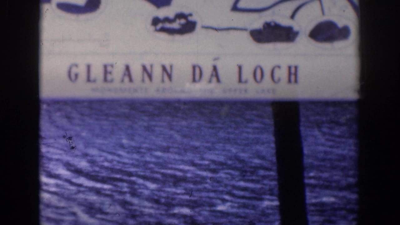 Gleann Da Loch sign above blue lake water with a dark vertical tree trunk.
