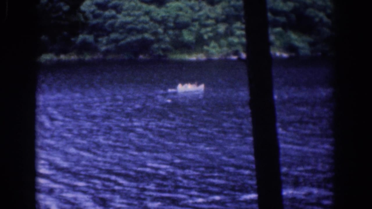 1961 8mm frame of a small boat moving across a blue lake, with wooded shoreline and tree silhouettes in the foreground.