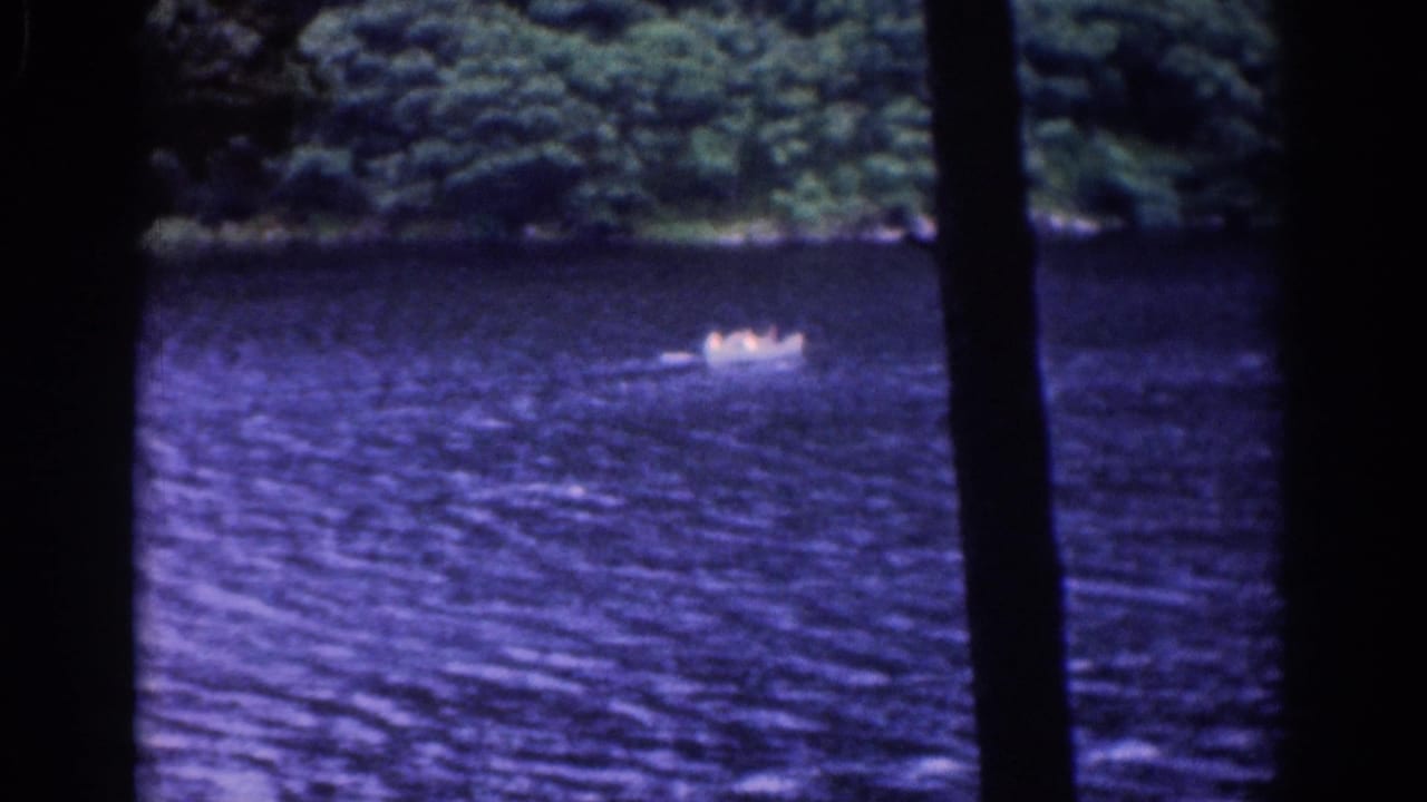 1961 8mm frame showing a distant small boat on the lake, framed by dark trees and a wooded far bank.