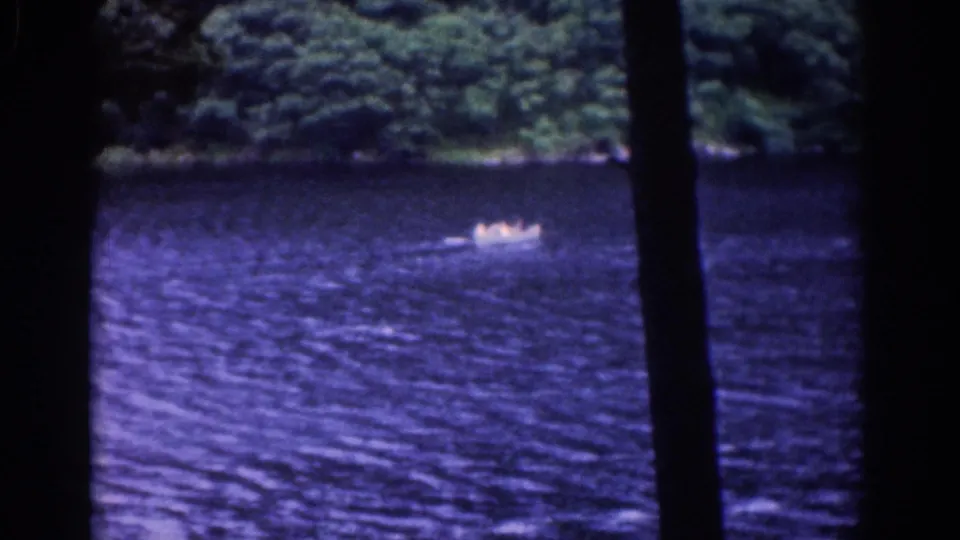 1961 8mm frame showing a distant small boat on the lake, framed by dark trees and a wooded far bank.