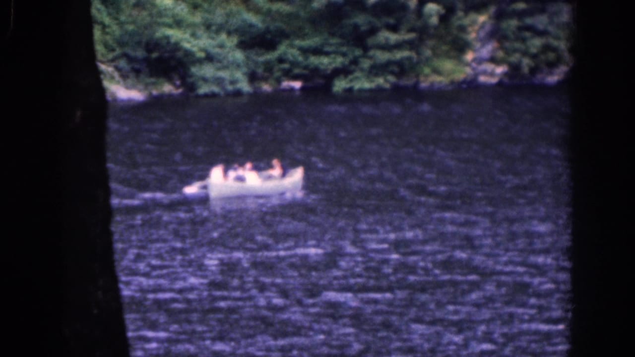 1961 Irish home-movie frame showing a white rowboat carrying several people across dark rippled water near a wooded shore.
