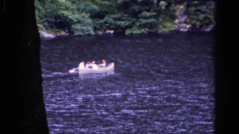 1961 Irish home-movie frame showing a white rowboat carrying several people across dark rippled water near a wooded shore.