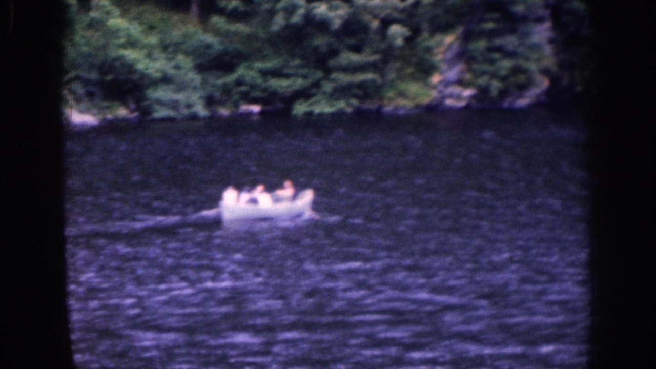1961 home-movie frame of visitors in a small white rowboat on a lake, with soft focus and dark tree framing.