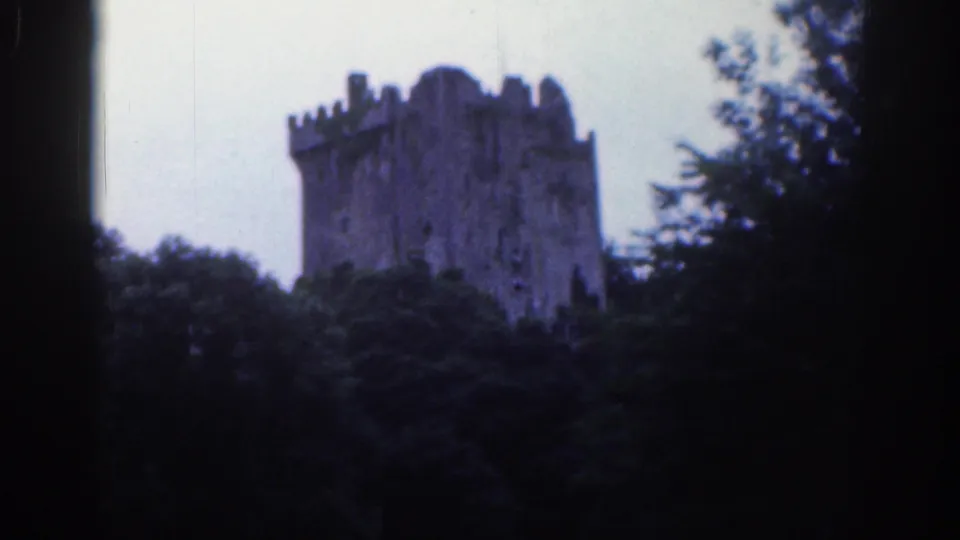 Distant 1961 home-movie view of Blarney Castle's stone tower above dense greenery, with soft focus and dark frame edges.