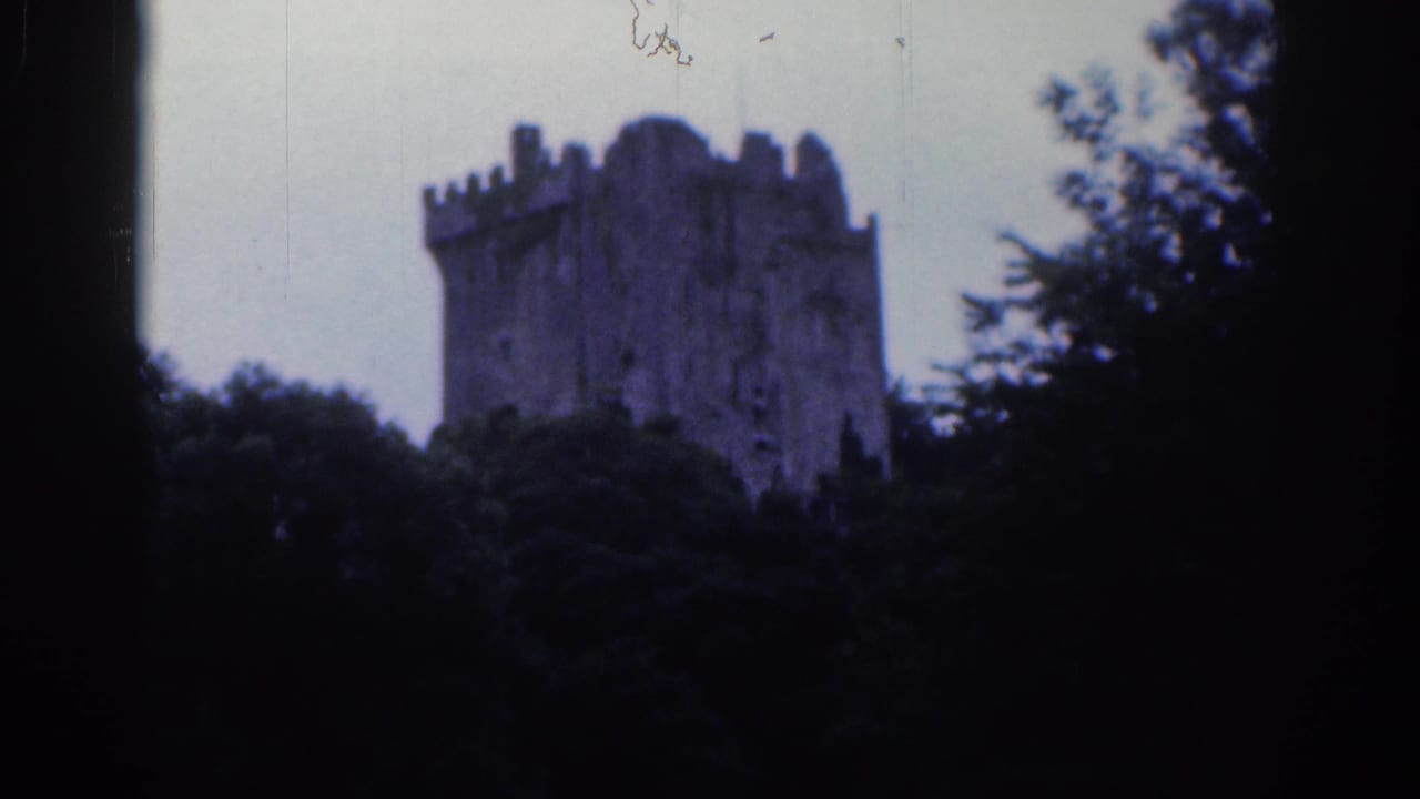 1961 Dublin home-movie frame showing a distant stone castle tower above dense trees, with dark 8mm frame edges.