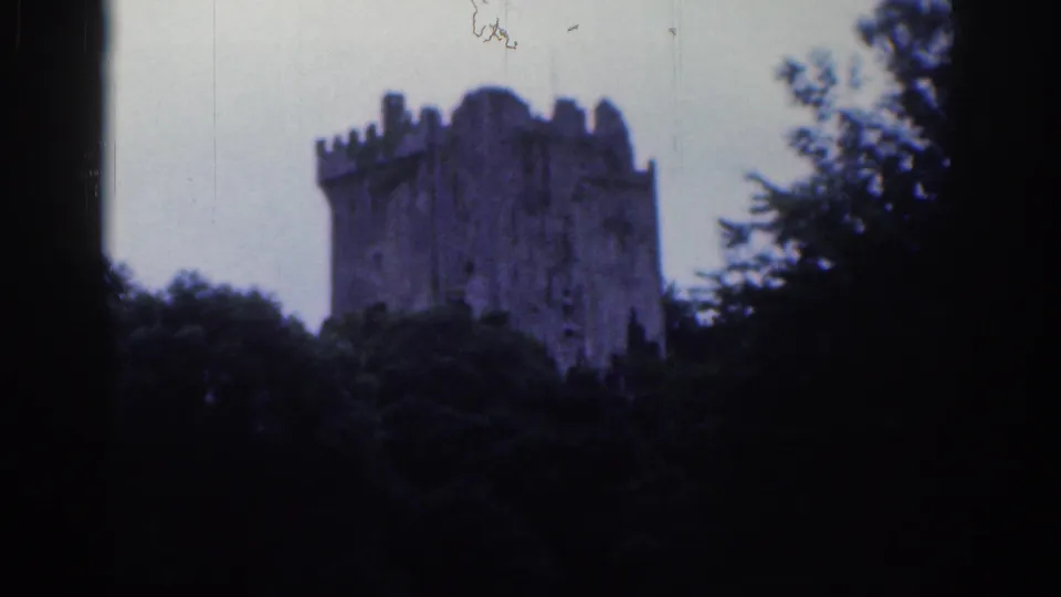 1961 Dublin home-movie frame showing a distant stone castle tower above dense trees, with dark 8mm frame edges.