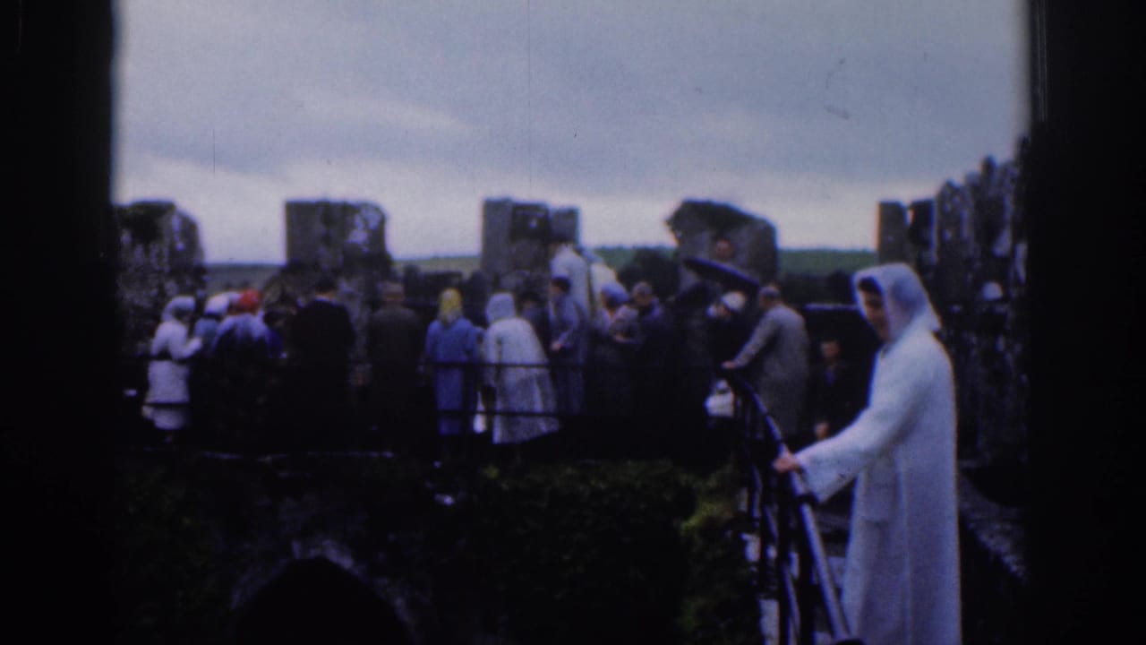 1961 Dublin home-movie footage shows tourists near ancient ruins, with a white-robed figure standing beside a railing in the foreground.