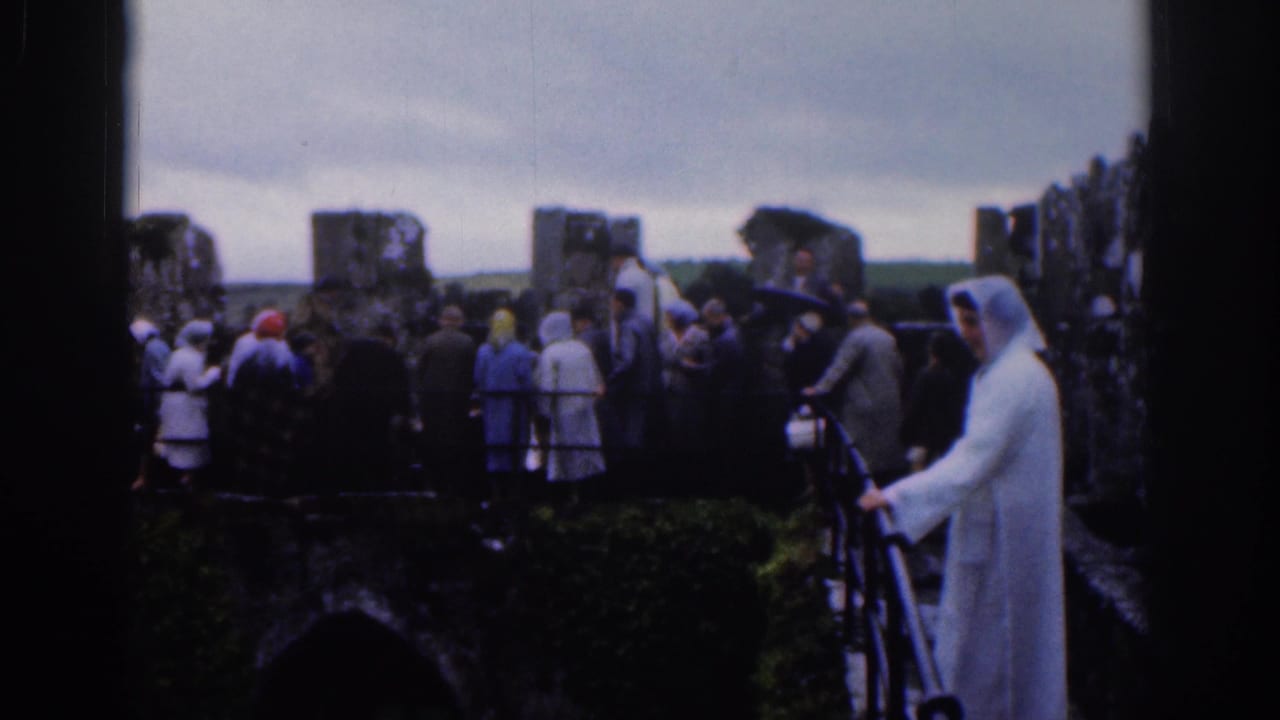 Tourists in mid-century clothing view stone ruins near Dublin in 1961, with a white-robed foreground figure and overcast sky.