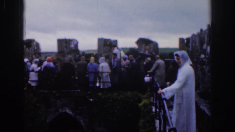 Tourists in mid-century clothing view stone ruins near Dublin in 1961, with a white-robed foreground figure and overcast sky.