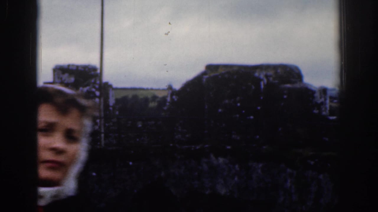 A woman's face enters the lower-left edge of a 1961 Dublin home-movie frame near rough stonework and distant fields.