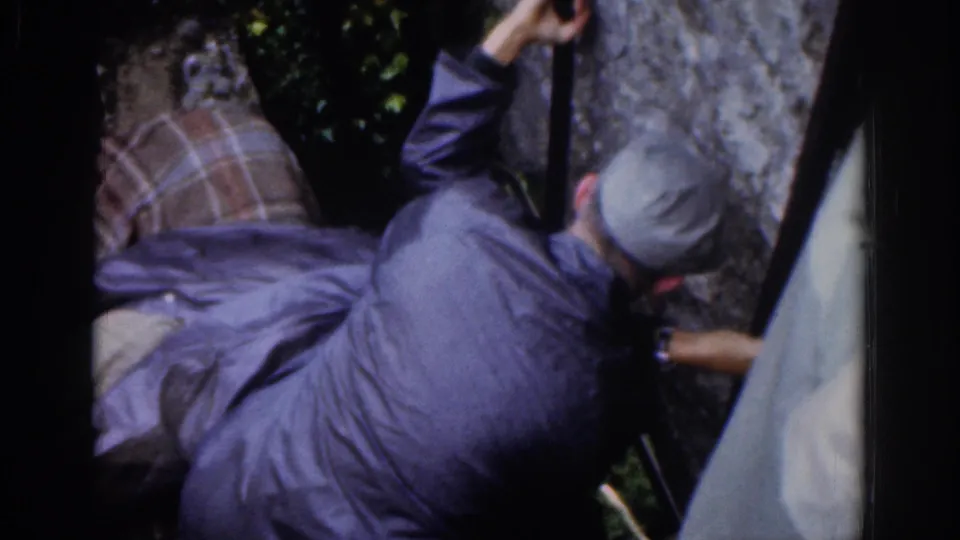 A man in a cap and dark jacket climbs or steadies himself on a steep stone wall in 1961 Dublin home-movie footage.