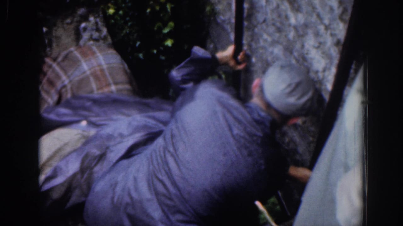 In Dublin home-movie footage from 1961, a man in a gray cap and dark jacket leans over another man near a stone wall and metal pole.