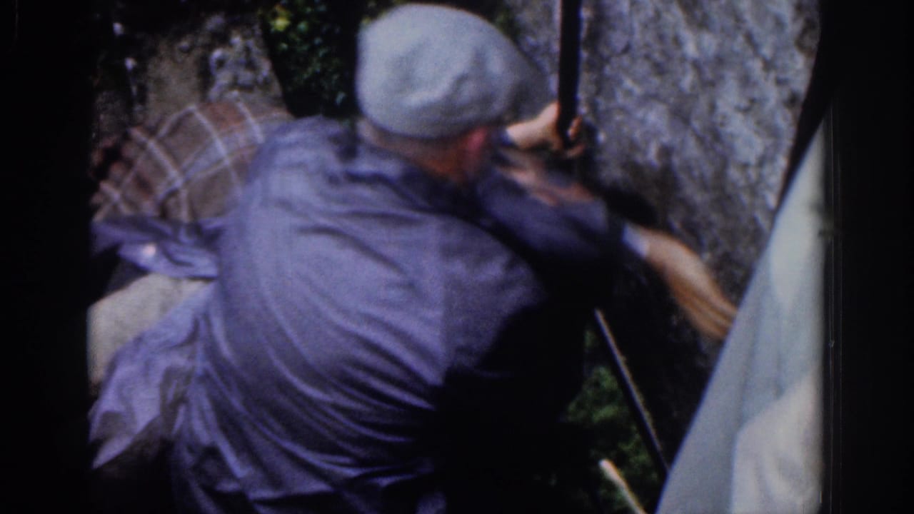 Man in gray cap pulls another man upward beside a stone wall.