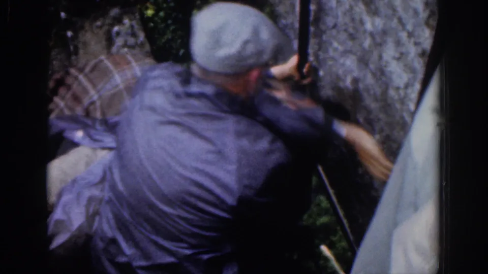 A 1961 Dublin frame shows the capped man straining near a stone wall while pulling or steadying another man in a plaid shirt.