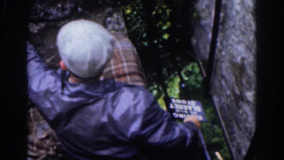 The final frame shows the capped man above the plaid-shirted man near greenery, a stone wall, metal pole, and the Blessing Stone sign.