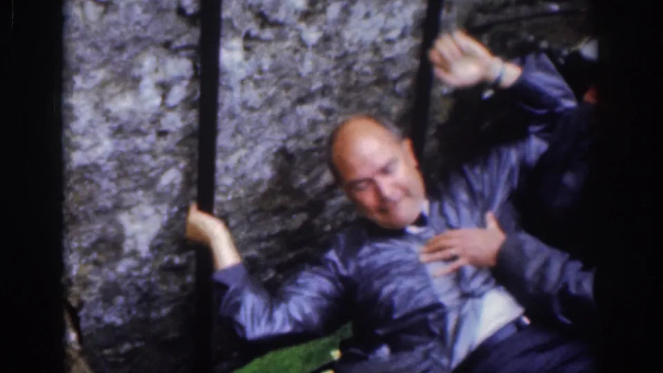 A suited adult visitor leans back from the Blarney Stone position, holding an iron bar as another person supports him.
