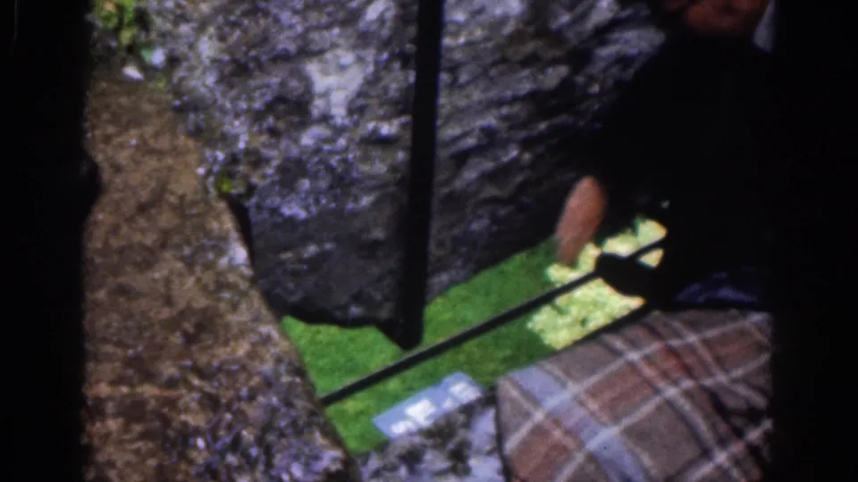 The camera shifts toward the stonework and bars, with grass below and part of a person or plaid garment at the edge.
