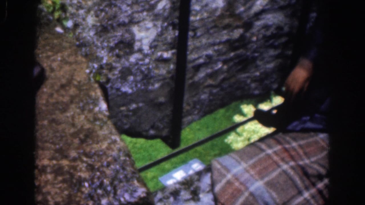 1961 Dublin home-movie frame looking past castle stone toward a nearby person’s plaid kilt or blanket and dark shoe above grass.