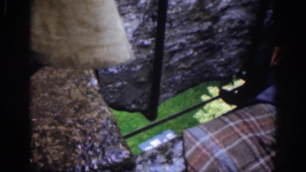 Rough stone opening with plaid fabric, dark shoe, green grass, and a small light item below.