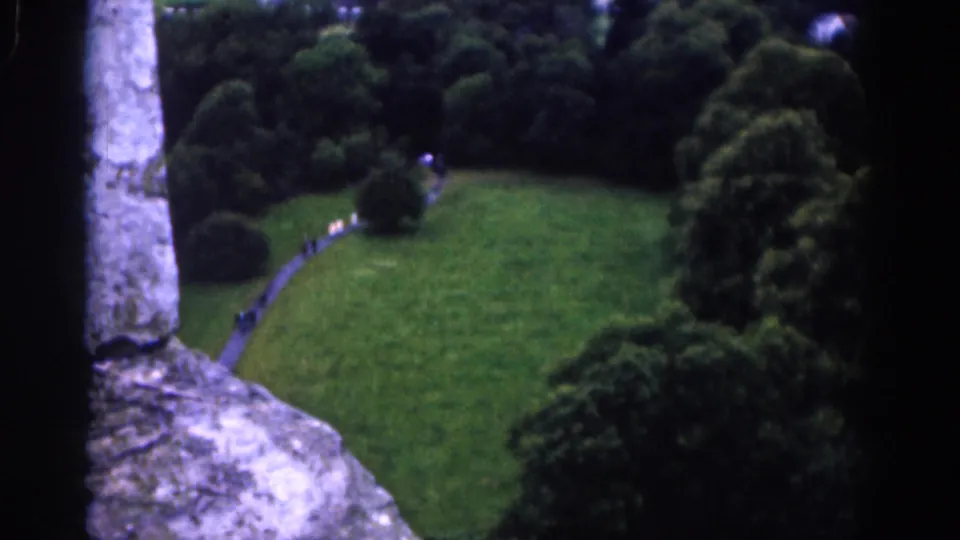 1961 Dublin home-movie frame looking through rough stonework toward a green park, winding path, dense trees, and hills beyond.
