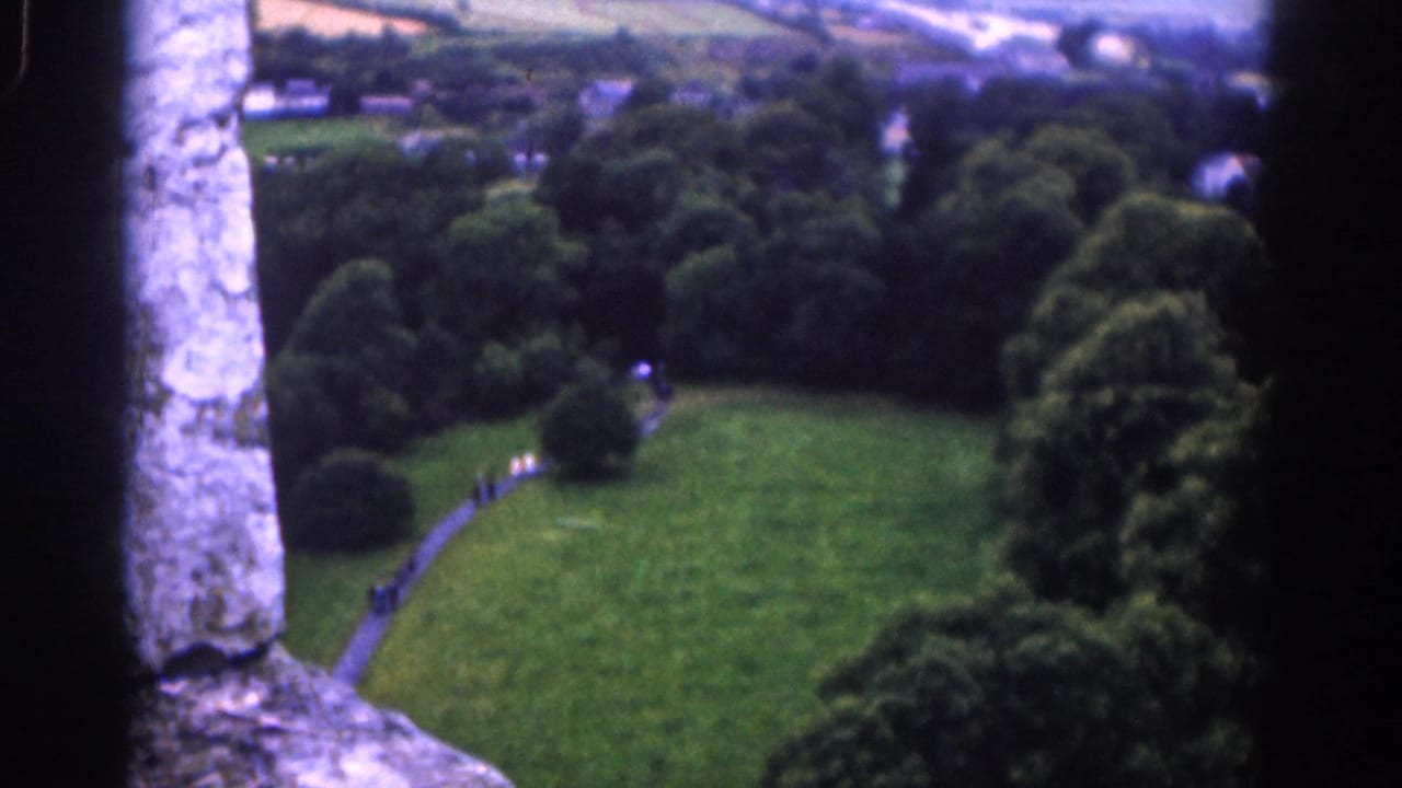 Wider elevated view of a park path, trees, fields, and distant buildings framed by stone edges.