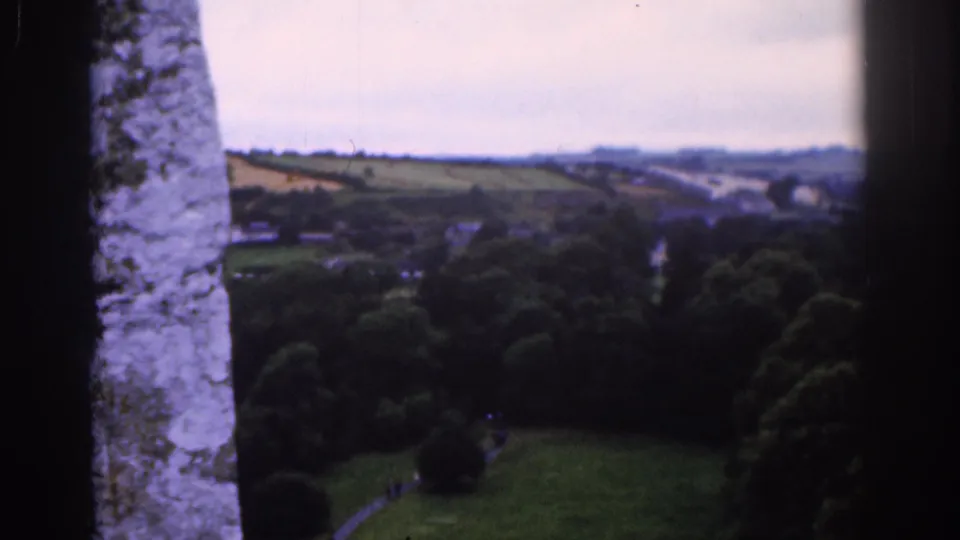 Final 1961 Dublin frame looking past rough stonework toward wooded grounds, open fields, a road or path, and a hazy horizon.