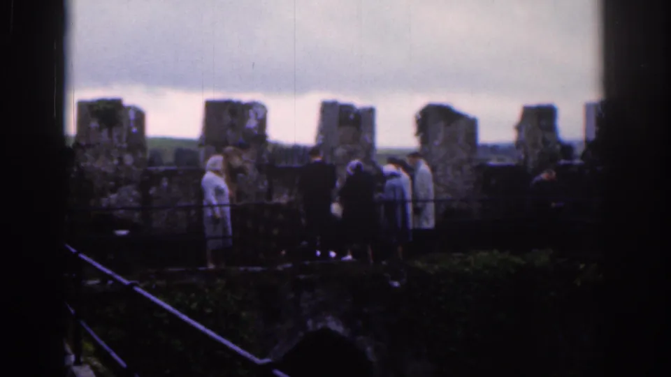 1961 Dublin home-movie frame showing visitors near old stone ruins, seen through dark foreground framing.