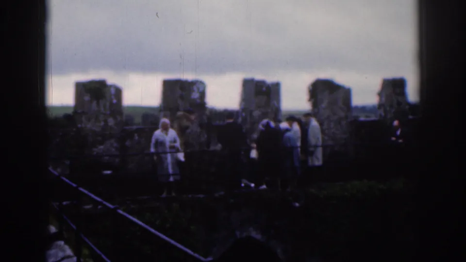 1961 Dublin travel footage frame with visitors beside ancient-looking stonework, distant hills, and heavy cloud cover.