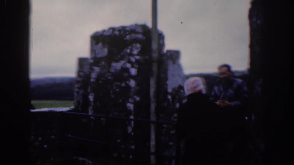 1961 Dublin home-movie frame showing adult visitors beside weathered stone ruins, railings, and distant open landscape.