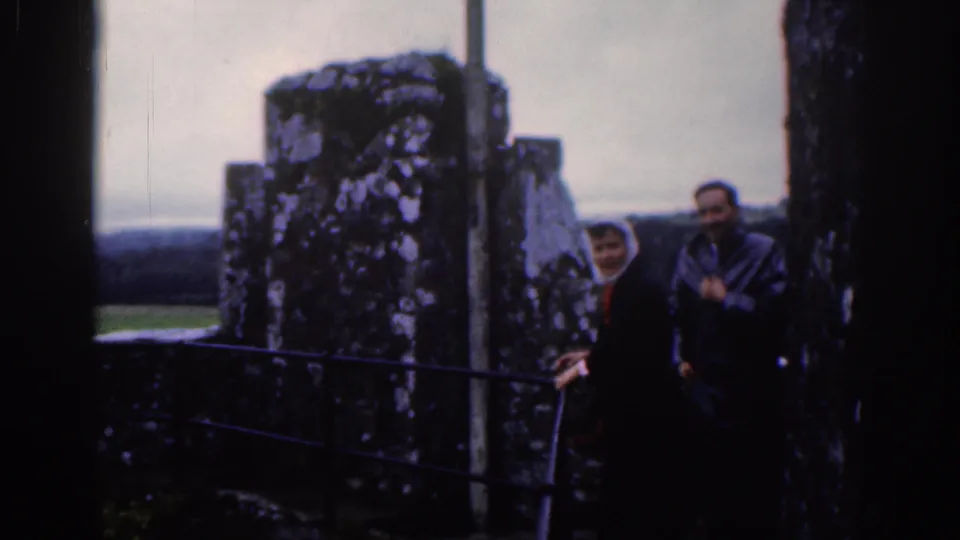 1961 Dublin frame with a woman in a white headscarf and a man beside dark stone ruins and metal railings.