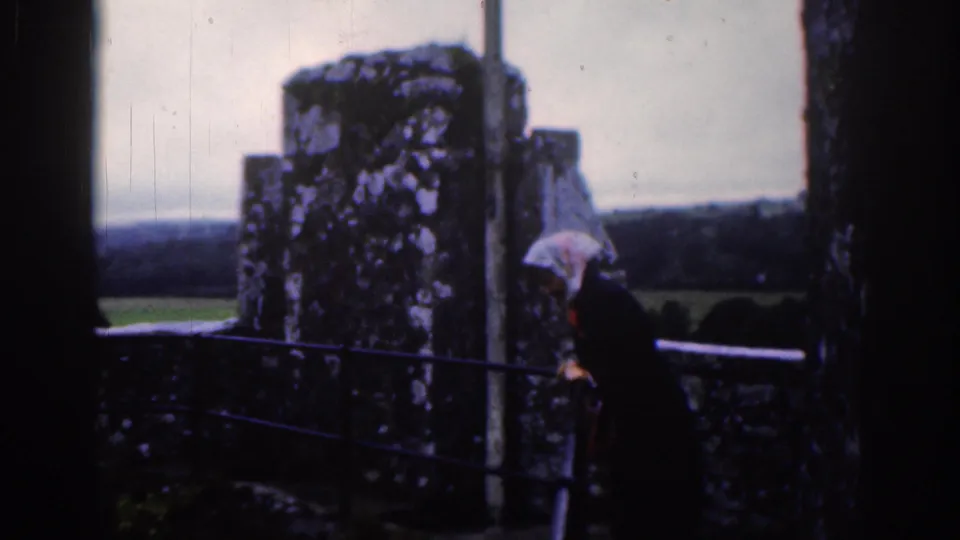 1961 Dublin home-movie frame showing a woman in a white headscarf moving along railings by weathered ruins.