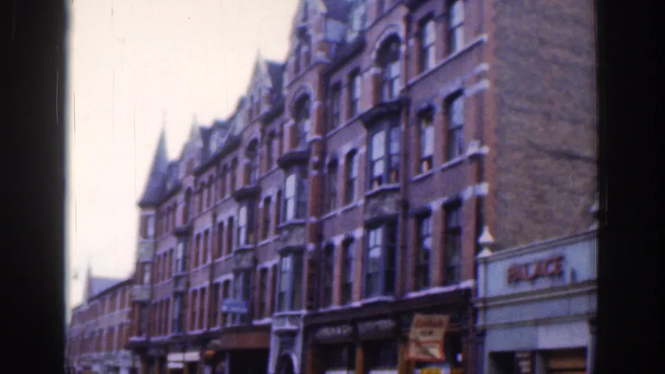 1961 Dublin street view showing ornate red brick Victorian-style buildings, shopfronts, and a pale sky in soft 8mm color.