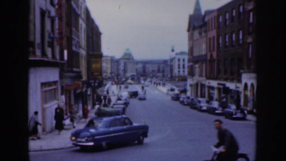 1961 Dublin street scene with a blue car driving past, parked cars, pedestrians on the sidewalk, and brick commercial buildings.