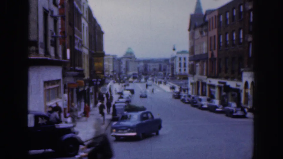 1961 Dublin street view with mid-century cars moving past pedestrians, storefronts, brick facades, and a distant domed building.