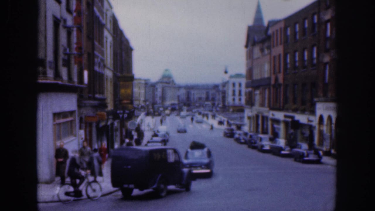 1961 Dublin street scene showing a dark utility van, parked mid-century cars, pedestrians near the curb, and brick commercial buildings.