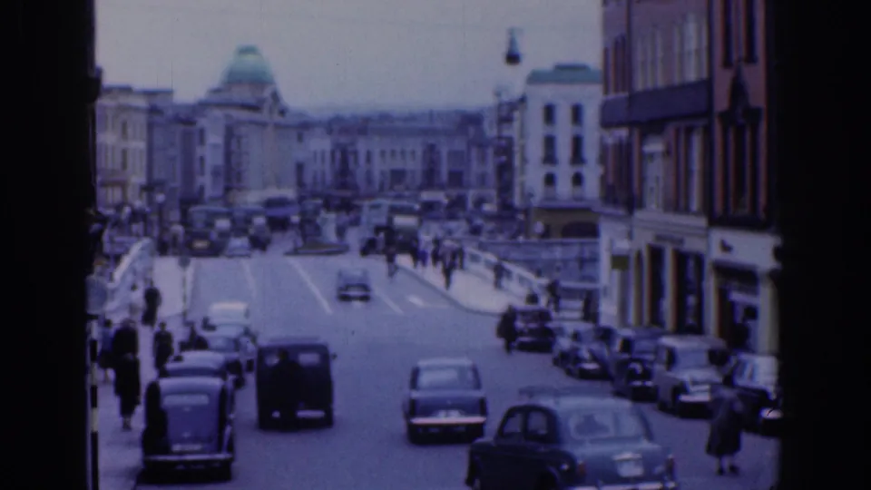 1961 Dublin street view with mid-century cars approaching the camera, pedestrians along both sidewalks, and a domed building beyond the traffic.