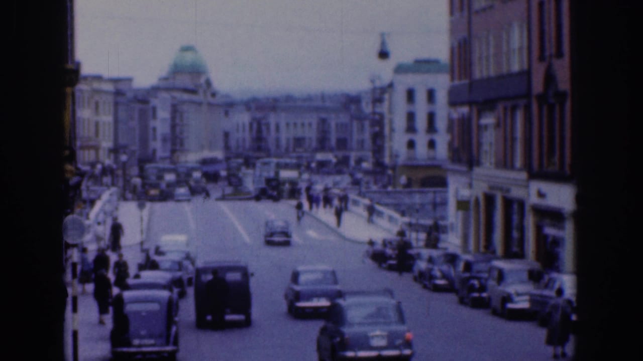 1961 Dublin street view with classic cars moving along a broad roadway, pedestrians on the sidewalks, masonry buildings, and a green-domed building in the distance.