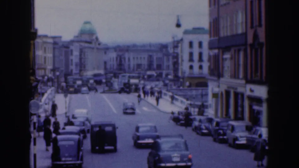 1961 Dublin street view with classic cars moving along a broad roadway, pedestrians on the sidewalks, masonry buildings, and a green-domed building in the distance.