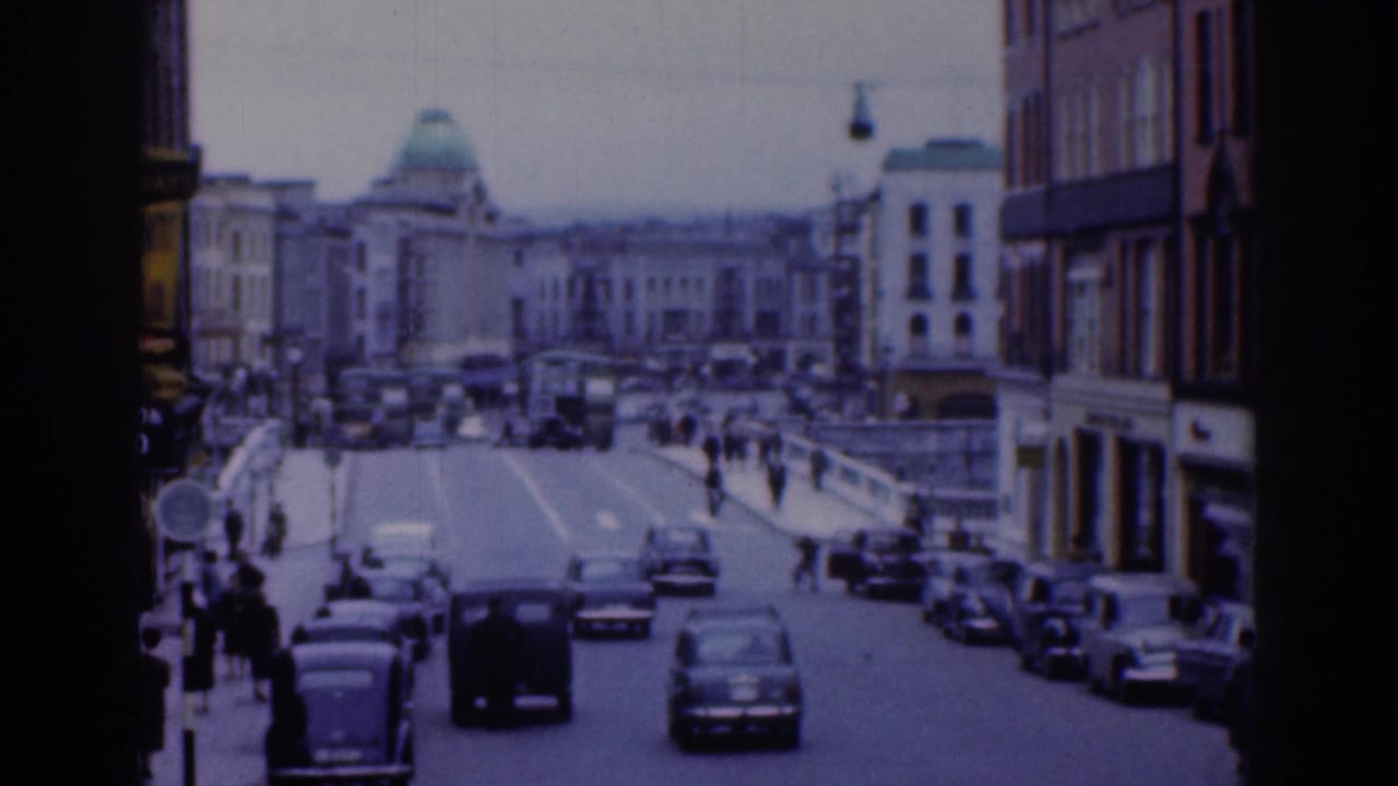 1961 Dublin city street with moving cars, parked vehicles along the curb, pedestrians near the roadway, and a domed building visible beyond the traffic.